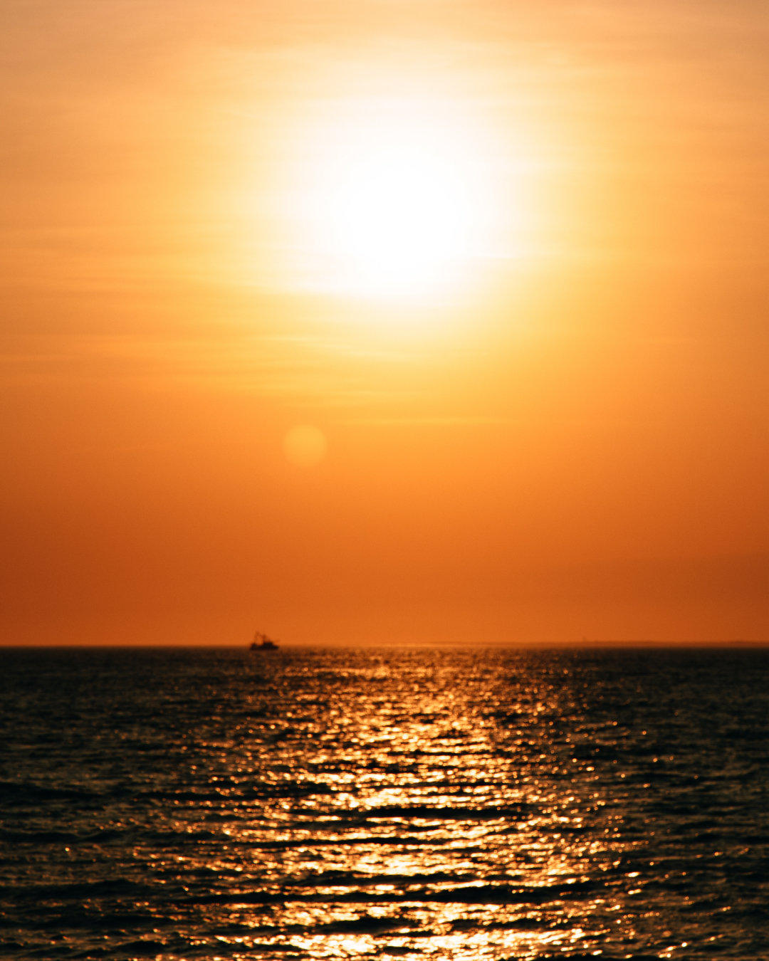 Sunset over a calm sea, with vibrant orange and yellow hues reflecting on the water, and a small silhouette of a boat in the distance.